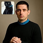 A sleek black cat with striking green eyes and a blue collar sits regally on a wooden surface, staring curiously at the camera with a plain wall and a power outlet in the background.