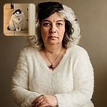 A fluffy white cat with striking black markings on its head sits patiently on a tiled floor near a doorway, looking slightly regal but also a bit unimpressed by the camera's attention.