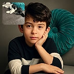 A fluffy black and white puppy lounges adorably on a textured gray couch next to a plush turquoise decorative pillow, looking up with big, soulful eyes that melt hearts instantly.