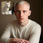 A charming white cat with striking blue eyes sits elegantly on a white surface, looking curiously at the camera. The background is simple and slightly blurred, highlighting the cat's soft fur and delicate whiskers. Nearby, a gold-capped item and a black spherical object add subtle detail to the scene.