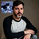 A cozy black and white cat lounges on a plaid blanket, looking intently at something off-camera. The background shows a wooden dresser and some colorful items, creating a warm indoor scene with a slightly soft focus.