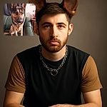 A charming German Shepherd sits attentively on a green leather couch, tongue slightly out, wearing a silver chain collar. The background shows a casual living space with a dark curtain and household items, adding a cozy and homey vibe.