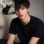 A curious black kitten with a tiny pink tongue peeking out sits attentively on a textured cream bedspread next to a sleek laptop, with soft natural light highlighting its shiny fur and big ears.