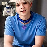 A fluffy white dog with a blue collar and a heart-shaped tag looks curiously at the camera while sitting on a textured blanket, with a cozy indoor background slightly out of focus.