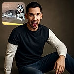 A majestic husky lounges comfortably on a sunlit concrete floor against a textured wall, tongue out and eyes sparkling with playful charm, looking like it just finished a fun outdoor adventure.