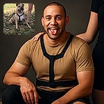 A happy dog with a black harness sits on a grassy patch covered with dry leaves, tongue out, looking content. A person's legs stand behind the dog, suggesting companionship and an outdoor walk on a sunny day.
