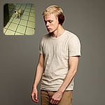 A small white dog with black spots stands on a tiled floor in a dimly lit room, looking slightly away from the camera as if pondering the mysteries of indoor life.