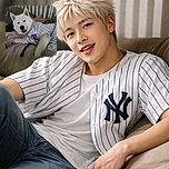 A fluffy white dog lounging comfortably on a bed, wearing a striped shirt that makes it look like it's ready for a baseball game, with a cozy and slightly cluttered background.