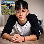 A charming black and white puppy lies stretched out on a grassy patch, basking in the sunlight with a curious and calm expression. The background shows a parking lot and a building under a clear blue sky, giving the scene a peaceful suburban vibe.