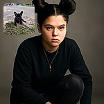 A serious-faced black French Bulldog puppy sits on a patch of dirt and grass on a quiet street with a colorful mural in the background, looking ready for its next adventure on a blue leash.
