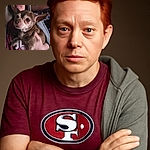 A close-up shot of a small brown Chihuahua being held by a person wearing a maroon San Francisco 49ers shirt and a grey hoodie. The dog looks curiously at the camera with big expressive eyes, while the person behind has a neutral expression. The background is dimly lit, adding a cozy indoor vibe.