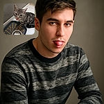A curious tabby cat with striking green eyes is caught mid-lick next to an empty metal food bowl on a polka-dotted mat, sitting on a tiled floor by a plain wall and wooden step.