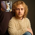 A fluffy cream-colored dog lounges regally on a wicker chair, looking like it owns the place, while a person in a cozy hoodie sits with their back to the camera, creating a cozy indoor scene with warm lighting and homey decor.