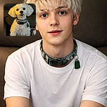 A fluffy white dog with curly fur sits attentively on a dark leather chair, wearing a collar with tags. The lighting casts a warm glow on the dog, highlighting its expressive eyes and soft fur. A cozy blanket with a playful pattern is partially visible beside the dog, adding a touch of homely comfort.