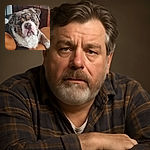 A close-up photo of a large, fluffy dog with a mix of brown, white, and gray fur resting its head on a tan leather couch, giving a soulful and slightly grumpy expression, with a wooden piece of furniture in the background.
