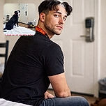A black and white dog sitting on a white bed looks back towards the camera with a curious expression, wearing a collar. The room has a wooden floor, a white door, a chair, a trash bin, and some dog toys scattered on the floor. Bold red text at the bottom reads 'BAMBAM IS MY ESA BOY'.