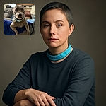 A curious gray dog with a blue collar leans over a dark blue blanket on a couch, staring directly into the camera with soulful eyes. The background shows a cozy living room with a TV, furniture, and soft lighting, slightly blurred to keep the focus on the dog's expressive face.