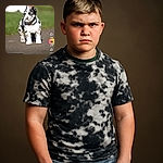 A chunky American Bulldog puppy with a striking black and white coat stands confidently on a paved path, looking curiously to the side. The background is a soft blur of green grass and out-of-focus objects, while the image is overlaid with social media interface elements including likes, comments, hashtags, and buttons.