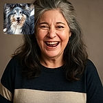 A fluffy, happy dog with a thick, multi-colored coat smiles joyfully at the camera, its bright eyes and open mouth radiating pure canine cheer against a natural outdoor background.