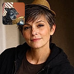 An adorable dog with a tiny straw hat perched on its head is being gently held by a person wearing a puffy jacket. The background features warm orange walls with sunlight casting striped shadows, giving the scene a cozy and playful vibe.