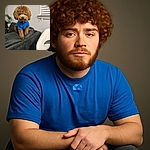 A fluffy brown dog wearing a cozy blue sweater sits perched on the back of a dark gray couch, looking adorably serious. The background shows a neutral wall with a window and blinds, alongside a white modern chair and a green plant adding a touch of life.