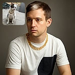 A regal white and black cat wearing a chunky gold chain sits on a tiled patio, looking off to the side with a calm and composed expression, surrounded by outdoor furniture and a wooden fence in the background.