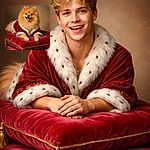 A fluffy Pomeranian sits like royalty on a plush red cushion, draped in a tiny velvet royal cloak with gold trim — tongue out, bright eyes, and enough pomp to demand a coronation (or at least extra treats).