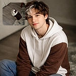 A fluffy brown-and-white dog sits half on a shaggy gray rug and half on a wood floor, looking up with big pleading eyes as if auditioning for the role of 'Chief Treat Officer'. A closet door and a small wooden side table with a cup sit in the background.