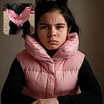 A fluffy black dog is lounging on a carpet, wearing a puffy pink jacket that looks comically oversized, giving off serious fashion model vibes while stretching out its front legs like it's ready for a cozy nap or a runway walk.