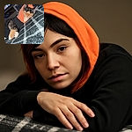 A cozy cat dressed in a tiny hoodie lounges comfortably on a soft, checkered blanket, looking slightly sleepy but undeniably adorable, with a TV and some household items blurred in the background.