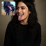 A fluffy black dog with a shiny coat stands outdoors on earthy ground, tongue playfully hanging out, looking off to the side with bright eyes and perked ears, capturing a joyful and lively moment in nature.