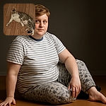 A relaxed tabby cat with white paws and belly lounges on a wooden floor, looking off to the side with a calm demeanor. The warm-toned floor contrasts with the cat's soft fur, creating a cozy indoor scene.