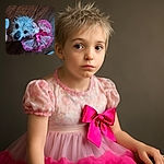 A scruffy little dog wearing a frilly pink dress with a big bow, looking up with curious eyes on a wooden floor. There's a hand gently holding the dog, and a foot with a sock visible at the edge, adding a cozy home vibe.