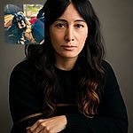 A fluffy black and tan dog lounges comfortably on a cozy couch next to a person, basking in warm sunlight streaming through a window. The dog's soulful eyes gaze towards the camera, framed by soft fur and a relaxed home setting with a blurred outdoor view in the background.