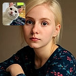 A curious white cat with big green eyes leans in close to the camera, its pink nose and whiskers prominently displayed. The background shows a cozy living room with a TV on and a patterned blanket in the foreground adding a splash of color.