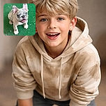 An adorable French Bulldog puppy with a speckled face looks up with big, curious eyes and a slightly open mouth, standing on a green textured surface with a pleasing bokeh background.