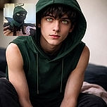 A charming black cat with striking green eyes is wearing a cozy green hoodie, sitting on a soft pink and black blanket with a curious and slightly mischievous expression, while a framed poster decorates the blurred background.