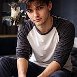 A curious tabby cat with white paws and chest is perched on a person's lap, gazing intently with a slightly tilted head, set against a cozy indoor background with dark curtains and a textured wall.