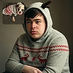 A cute bulldog puppy dressed in a festive sweater and a cozy hat with antlers, sitting on a textured carpet and giving a slightly grumpy but adorable look.