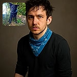 A wet black dog with a blue bandana stands alert in a lush green forest, looking curiously at the camera with ears perked up, surrounded by vibrant foliage and soft natural light.