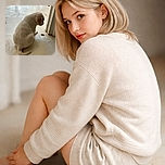 A small, light-colored puppy sits quietly on a tiled floor near a doorway, seemingly focused on a small dark object in front of it. The soft lighting highlights the puppy's fluffy fur and the clean, marble-like tiles add a calm, indoor atmosphere.