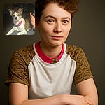 A small dog with a pink collar lounges comfortably on a textured surface, looking directly at the camera with curious eyes. The background is dimly lit with a brick wall and a hand holding a phone visible on the right side, adding a cozy indoor vibe.