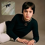 A slightly blurry photo of a black and white cat lounging on a wooden floor, looking curiously at the camera with a collar tag shining.