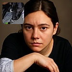 A close-up photo of a black dog with soulful brown eyes resting its paw on a gray surface, surrounded by a cozy blanket and a green-black object, with a human arm partially visible nearby.
