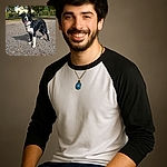 A joyful black and white dog stands alert on a sunlit gravel path, tongue out and ears perked, with a leafy tree and suburban houses in the background, radiating pure canine happiness.