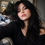 A close-up selfie of a fluffy black cat with striking green eyes lounging on a cozy bed with patterned sheets, looking slightly sleepy or uninterested.