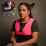 A small dog with a glowing pink collar sits attentively on a black office chair, perched atop a crumpled piece of cloth. The cozy indoor setting features a patterned rug and wooden paneling in the background, giving off a warm, lived-in vibe.