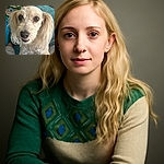 A close-up photo of a small, curly-haired white dog with large expressive eyes and slightly open mouth, sitting comfortably on a blue couch with colorful cushions in the background. The dog's fur looks soft and slightly tousled, giving it a cute and friendly appearance.
