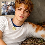 A relaxed orange and white cat lounges outdoors on the ground, basking in dappled sunlight with leaves and grass around, wearing a blue collar with a heart-shaped tag.
