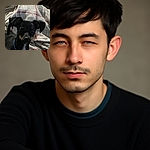 A slightly blurry photo of a small black dog with tan markings wearing a blue collar, sitting or lying in front of a cozy cream-colored blanket with red stripes. The dog looks a bit sleepy or relaxed in a warm indoor setting.